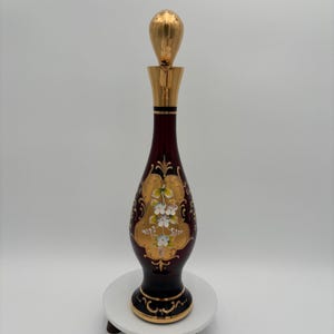 May include: A decorative glass bottle with a gold stopper. The bottle is a deep burgundy colour, featuring gold floral designs and accents. The base and neck are also trimmed in gold.