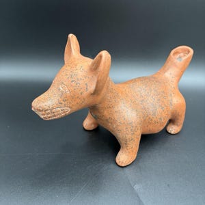 Pode incluir: Estatueta de cão em cerâmica cor de terracota com superfície salpicada. O cão tem orelhas direitas, um focinho detalhado e um pequeno orifício na ponta da cauda. A estatueta é um objeto decorativo.