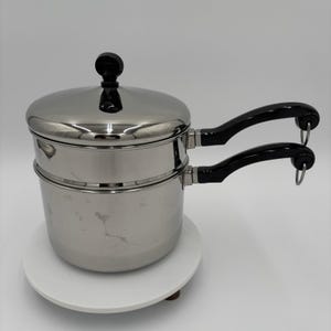 May include: Stainless steel double boiler with a lid and two black handles. The pot sits on a white, round surface. The lid has a black knob. The handles have a curved design.