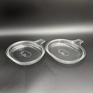 Może przedstawiać: Dwa przezroczyste szklane naczynia do pieczenia Pyrex z uchwytem z jednej strony. Naczynia mają owalny kształt z lekko podniesioną krawędzią. Naczynia znajdują się na ciemnej powierzchni.