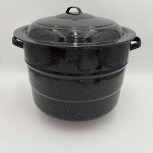Puede incluir: Una olla de conserva de esmalte negro con tapa. La olla tiene un patrón blanco moteado y dos asas laterales. La tapa tiene un pequeño asa en la parte superior. La olla está diseñada para conservar alimentos.