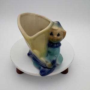 May include: A vintage ceramic planter in the shape of a pixie or elf. The planter is primarily yellow and blue, with a beige interior. The pixie figure has a smiling face and is wearing a blue outfit. The planter is on a white, round stand.