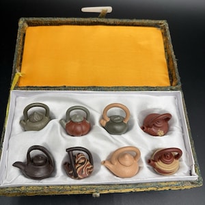 May include: A set of eight miniature ceramic teapots in a decorative box. The teapots are in various colors, including brown, green, and tan. The box has a yellow interior and a green and gold exterior.