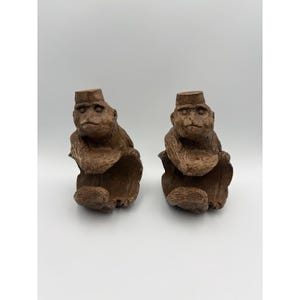 May include: A pair of brown, carved wooden monkey figurines, each wearing a small flat-topped hat. The monkeys are depicted in a seated position with their arms resting on their knees, featuring textured surfaces and detailed facial expressions.
