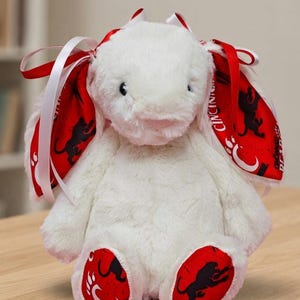 May include: A white plush rabbit toy with long ears lined with red fabric featuring black bear silhouettes and white paw prints. The rabbit has white and red ribbons tied at the base of its ears and matching red fabric on the soles of its feet.