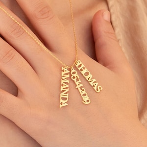May include: A gold-colored necklace with three vertical pendants. The pendants are engraved with the names "AMANDA" and "THOMAS", and the date "05.14.2019". The necklace is worn around the neck.