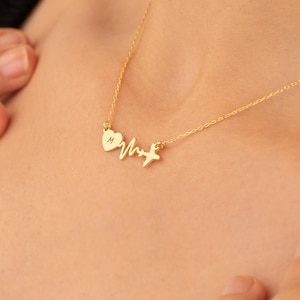 May include: A delicate gold necklace featuring a heart, a heartbeat line, and a star pendant. The necklace is worn around the neck and rests on the chest. The chain is thin and the pendant is small, creating a minimalist aesthetic.