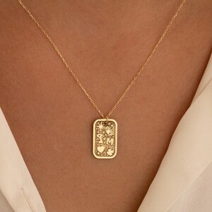Pode incluir: Um colar com pingente retangular dourado. O pingente apresenta desenhos gravados de sol, coração, trevo e iniciais. O colar é usado sobre uma camisa branca.