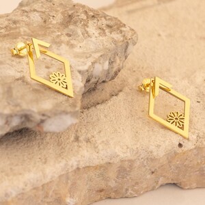 Puede incluir: Un par de pendientes dorados en forma de diamante. Cada pendiente presenta un diseño floral dentro de la forma de diamante. Los pendientes se muestran sobre una superficie de piedra clara y texturizada.