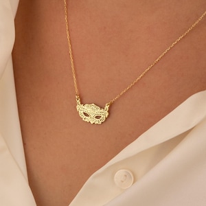 May include: A gold-colored necklace featuring a detailed mask pendant. The delicate chain and pendant are displayed against a cream-colored button-down shirt. The mask design has intricate cut-out details.