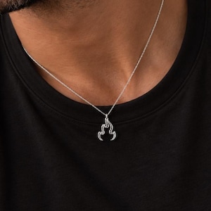 May include: A silver-toned necklace featuring a small, stylized flame-shaped pendant suspended from a delicate chain. The pendant has a polished metallic finish with a cutout design that mimics the flickering movement of fire, resting against a black shirt.