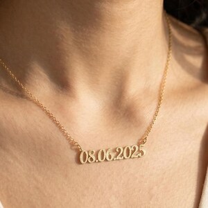 May include: A gold-toned metal necklace featuring a delicate chain and a central pendant displaying the numerical date 08.06.2025 in a serif font. The necklace rests against the neck, showcasing a polished finish and a simple, refined design.
