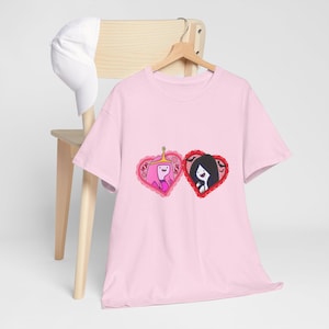 Pode incluir: Uma t-shirt de manga curta cor-de-rosa claro com uma impressão gráfica de duas molduras em forma de coração. Um coração contém uma personagem de cabelo cor-de-rosa com uma coroa dourada, e o outro uma personagem de cabelo preto rodeada de morcegos.