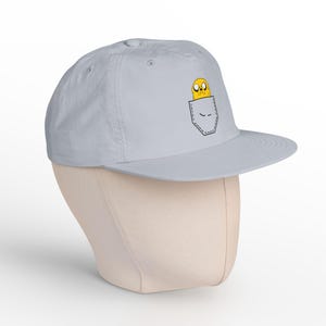 Könnte beinhalten: Hellgraue Baseballkappe mit einem Cartoon-Charakter in einem Taschen-Design. Der Charakter ist gelb mit schwarzen Umrissen. Die Kappe hat einen flachen Schirm und eine runde Krone, geeignet für den lässigen Gebrauch.