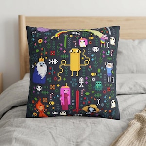 May include: A square throw pillow with a dark charcoal background featuring a colorful pixel art pattern of various cartoon characters, including a yellow dog, a blue-clad human, a pink-haired figure, a white-bearded king, and small floral motifs.