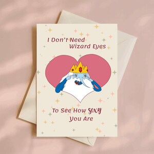 Op de afbeelding: Een wenskaart met een cartoonfiguur met een kroon en blauwe armen, tegen een roze hart. De kaart leest "I Don't Need Wizard Eyes To See How Sexy You Are" op een beige achtergrond.
