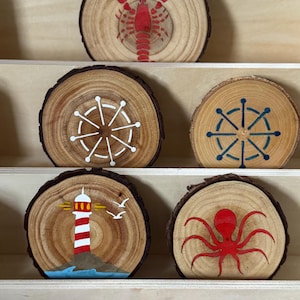 Puede incluir: Un conjunto de posavasos de madera con diseños náuticos. Los posavasos presentan una langosta roja, timones, un faro y un pulpo rojo. Cada posavasos es una rodaja de madera circular con borde de corteza natural.