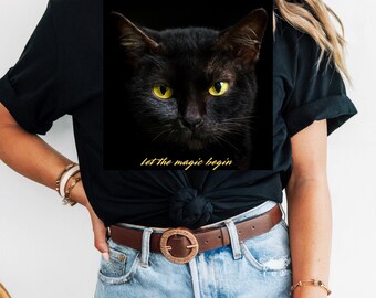 May include: Black t-shirt with a black cat image and yellow eyes. The text "let the magic begin" is printed in gold. The shirt is worn with blue jeans and a brown belt. The overall theme is a mystical cat.