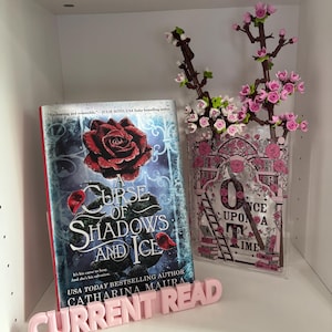 May include: A book titled "A Curse of Shadows and Ice" with a red rose on the cover, next to a decorative vase with pink flowers. A pink sign that says "CURRENT READ" is in front of the book.