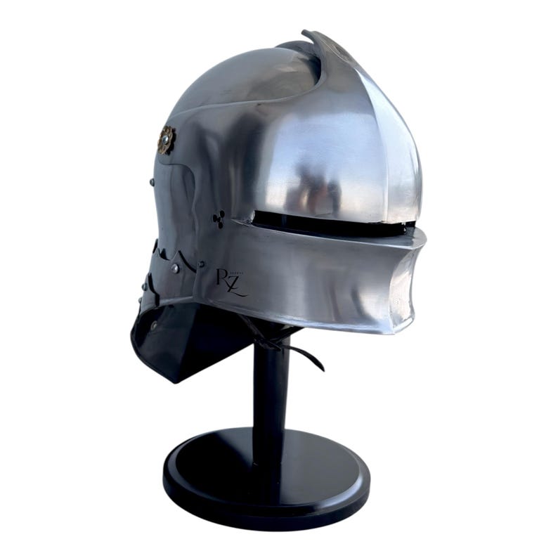 May include: A polished silver-colored metal helmet with a rounded dome, a horizontal visor slit, and a flared neck guard. The side features a small circular brass-toned emblem and the letters PZ engraved on the lower cheek section of the helmet.