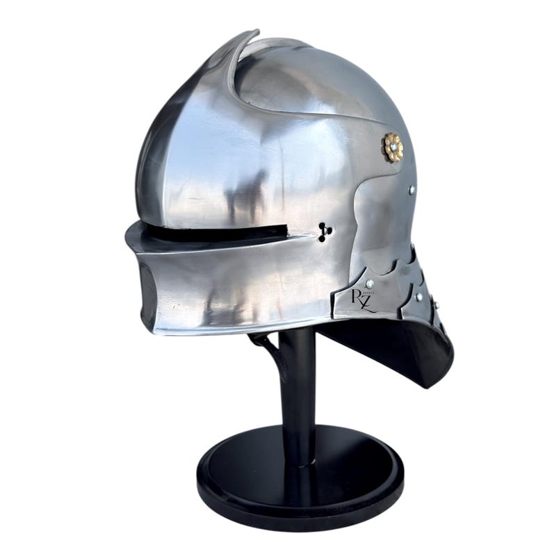May include: A polished silver-colored metal helmet with a rounded crown, a horizontal eye slit, and a flared neck guard. The helmet features a small gold-toned floral rivet on the side and the letters RZ printed in black on the lower right side.