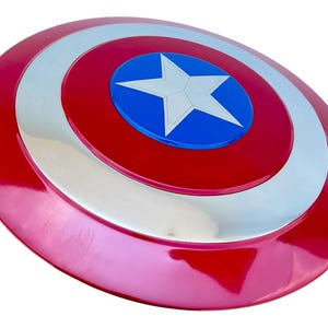 May include: A red, white, and blue shield with a central white star on a blue circle. The shield has a glossy finish and a circular design, reminiscent of a superhero's emblem.
