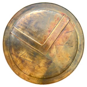 May include: A round, antique-style shield with a bronze finish. The shield features a prominent V-shaped design and a series of small, evenly spaced rivets. The overall aesthetic suggests a historical or fantasy theme.