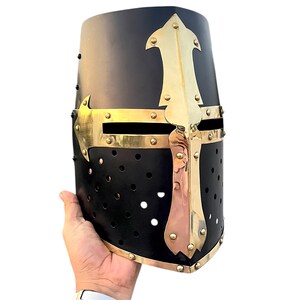 Pu&ograve; includere: Elmo medievale nero con croce e dettagli in ottone. L'elmo ha una parte superiore arrotondata ed &egrave; adornato da una prominente croce in ottone sul davanti. Sono visibili piccoli fori di ventilazione. Un pezzo storico o in costume.