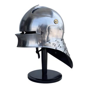 May include: A polished steel sallet helmet featuring a rounded crown, a horizontal eye slit, and a flared neck guard. The helmet includes a decorative gold-toned floral rivet on the side and the letters RZ printed in black on the lower side plate.