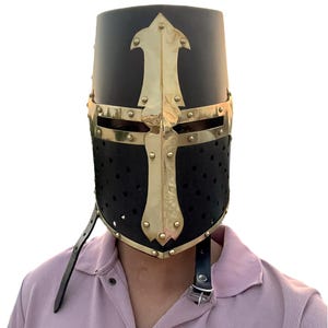 Pu&ograve; includere: Un elmo da crociato medievale nero e oro. L'elmo presenta una parte superiore cilindrica con un disegno a croce dorata e una protezione per il viso perforata. &Egrave; fissato con un cinturino in pelle nera.