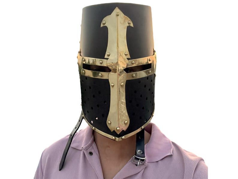 Pu&ograve; includere: Un elmo da crociato medievale nero e oro. L'elmo presenta una parte superiore cilindrica con un disegno a croce dorata e una protezione per il viso perforata. &Egrave; fissato con un cinturino in pelle nera.
