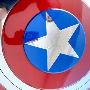 Peut inclure: Gros plan sur un bouclier aux couleurs rouge, blanc et bleu. Le bouclier pr&eacute;sente des cercles concentriques avec une &eacute;toile argent&eacute;e au centre d'un cercle bleu. Le bouclier est une r&eacute;plique d'un bouclier de super-h&eacute;ros.