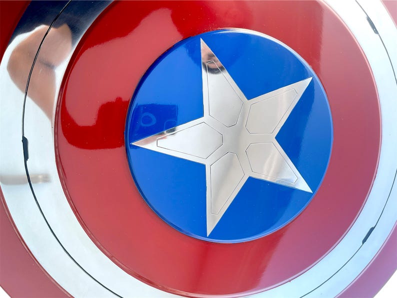 Peut inclure: Gros plan sur un bouclier aux couleurs rouge, blanc et bleu. Le bouclier pr&eacute;sente des cercles concentriques avec une &eacute;toile argent&eacute;e au centre d'un cercle bleu. Le bouclier est une r&eacute;plique d'un bouclier de super-h&eacute;ros.