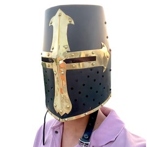 Pu&ograve; includere: Elmo nero da crociato medievale con un design a croce dorata. L'elmo presenta una parte superiore arrotondata, fori di ventilazione e un sottogola in pelle. La croce dorata &egrave; rivettata al metallo nero.