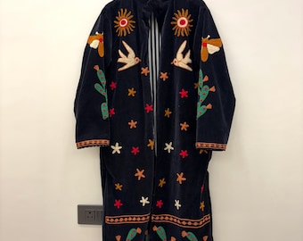 May include: A dark navy blue long coat featuring colorful embroidery of suns, birds, moths, and cacti. The garment has a high collar, long sleeves with orange-trimmed cuffs, and a black and white vertical striped lining visible at the front opening.