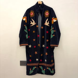 May include: A dark blue embroidered coat with a high collar. The coat features colorful embroidered designs of suns, birds, cacti, and stars. The coat has a long, flowing design and is hanging on a wooden hanger.