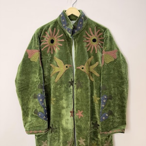 May include: An olive green velvet coat featuring embroidered suns, birds, and star motifs in shades of pink, yellow, and blue. The coat has a high collar, long sleeves, and a front opening, with a striped lining visible at the neckline.