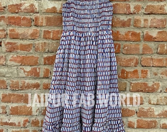 May include: A blue and white patterned dress with a ruffled neckline and hem, hanging on a wooden hanger. The dress features a repeating vertical stripe design in blue and white with red accents. The text "JAIPUR FAB WORLD" is printed on the dress.