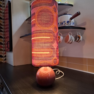 Könnte beinhalten: Eine zylindrische Lampe im Retro-Design mit einem rosa, orangefarbenen und gelben Muster. Vor der größeren Lampe befindet sich eine kleine, runde, rosa Lampe. Die Lampen stehen auf einer dunklen Oberfläche. Der Hintergrund umfasst Regale und Küchenutensilien.