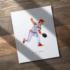 May include: A print featuring a baseball player in a Cincinnati Reds uniform, mid-pitch. The player is wearing a red cap, white jersey with "Cincinnati" across the chest, and red and white pants. The artwork is on a white background with a wooden surface.