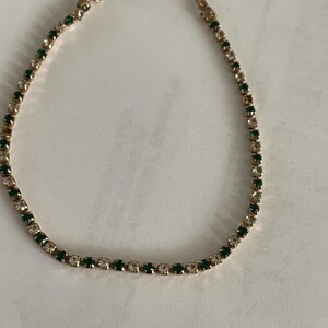 May include: A gold-toned bracelet featuring alternating green and clear gemstones. The bracelet has a delicate design with a clasp closure. The gemstones are small and evenly spaced along the length of the bracelet.
