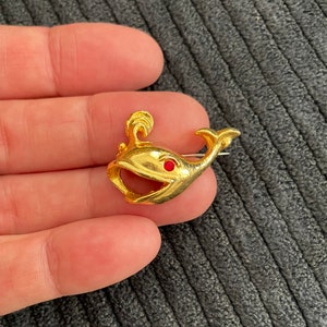 May include: A gold-toned whale brooch with a red rhinestone eye. The whale has a fin on its back and a curled tail.