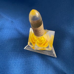 May include: A vintage perfume bottle with a yellow rose design inside a clear, triangular base. The bottle has a clear, rounded top and a label with text. The base is on a blue surface.