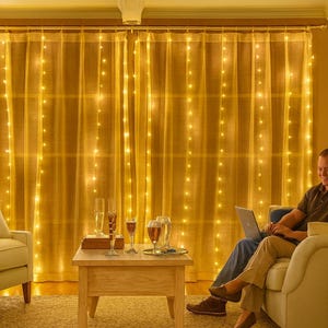 Puede incluir: Una acogedora sala de estar con una cortina transparente adornada con luces de cadena. Una pareja está sentada en un sofá crema, usando una computadora portátil. Copas de champán y una pequeña mesa contribuyen al ambiente.