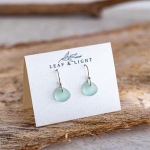 K&ouml;nnte beinhalten: Ein Paar Ohrh&auml;nger mit kleinen, glatten, mattierten hellgr&uuml;nen Meerglasst&uuml;cken, die an silberfarbenen Metallhaken h&auml;ngen. Die Ohrringe sind an einer wei&szlig;en Karte befestigt, die mit dem Text LEAF & LIGHT und einem Blattsymbol bedruckt ist.