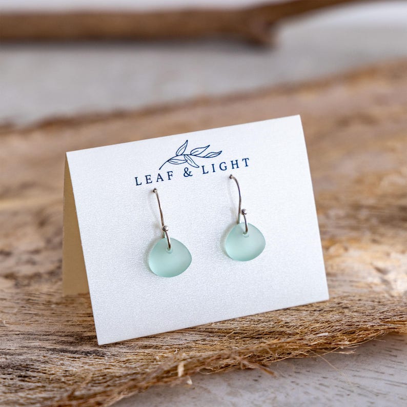K&ouml;nnte beinhalten: Ein Paar Ohrh&auml;nger mit kleinen, glatten, mattierten hellgr&uuml;nen Meerglasst&uuml;cken, die an silberfarbenen Metallhaken h&auml;ngen. Die Ohrringe sind an einer wei&szlig;en Karte befestigt, die mit dem Text LEAF & LIGHT und einem Blattsymbol bedruckt ist.