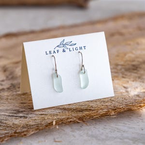 K&ouml;nnte beinhalten: Ein Paar Ohrh&auml;nger aus blass-seegr&uuml;nem Glas mit silberfarbenen Metallhaken, pr&auml;sentiert auf einer wei&szlig;en rechteckigen Karte mit dem dunkelblauen Schriftzug LEAF & LIGHT und einer kleinen Blattillustration &uuml;ber dem Markennamen.