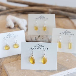 Könnte beinhalten: Vier Paar Ohrringe aus gelbem Meerglas mit Silberhaken, jeweils auf einer weißen Karte mit dem Aufdruck LEAF & LIGHT präsentiert. Die Ohrringe bestehen aus glatten, durchscheinenden gelben Glasstücken, die an einfachen Silberhaken hängen.