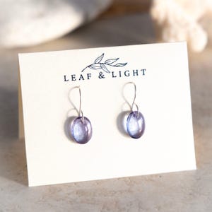 Könnte beinhalten: Ein Paar kleine, oval geformte, durchscheinende violette Glas-Ohrhänger mit silberfarbenen Ohrhaken. Die Ohrringe sind auf einer cremefarbenen Karte mit dem Text LEAF & LIGHT und einer kleinen Blattillustration über dem Markennamen befestigt.