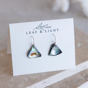 May include: A pair of dangle earrings featuring iridescent, triangular-shaped shell charms with dark edges and silver-toned hooks. The earrings are displayed on a white card printed with the text LEAF & LIGHT and a small leaf illustration.